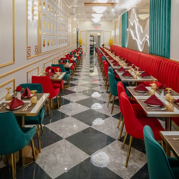 Restaurant interior with long rows of tables, red and teal chairs, and a black and white checkered floor.