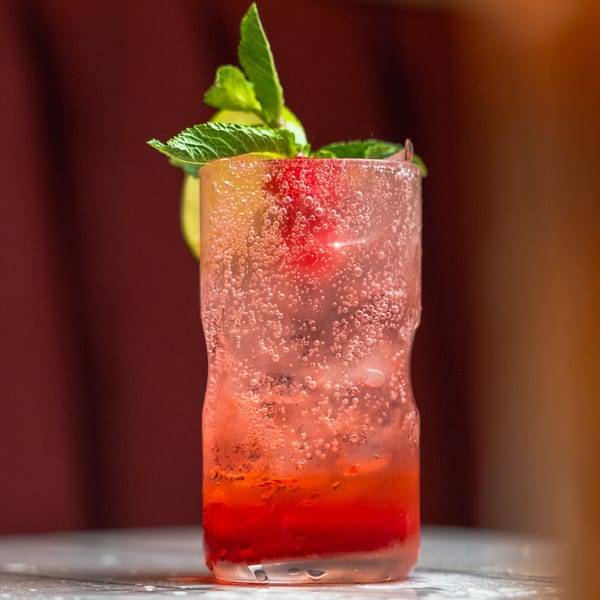 A vibrant red and pink bubbly drink, garnished with lime and mint, sits on a table.
