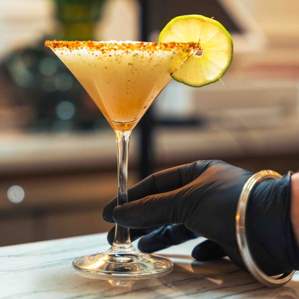 A hand in a black glove holds a martini glass with a spiced rim and lime garnish.