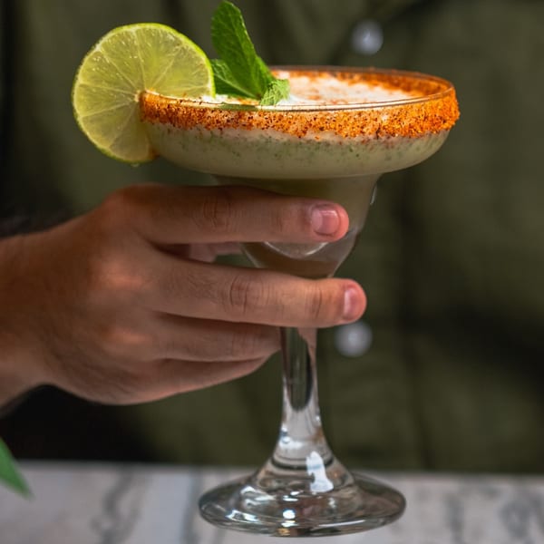 A hand holds a Red Chilli margarita, garnished with a lime slice, mint, and chili-salt rim.