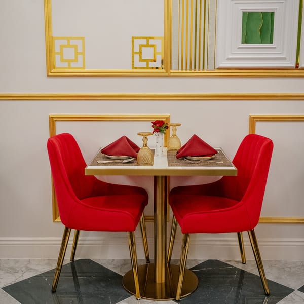 A dining table set for two with red chairs, gold accents, and a small vase of red flowers.