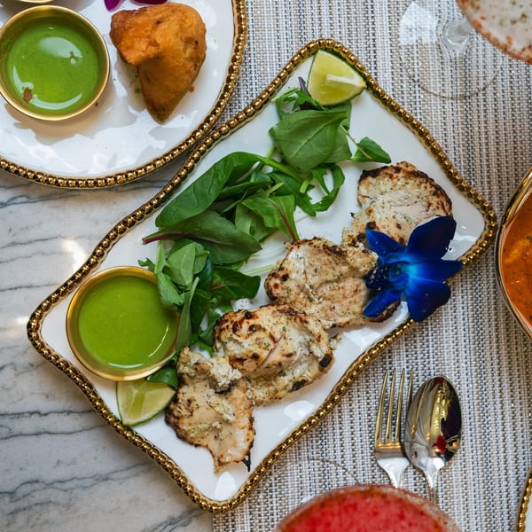 Indian grilled chicken with green chutney, lime, and a blue flower garnish on a white and gold plate.