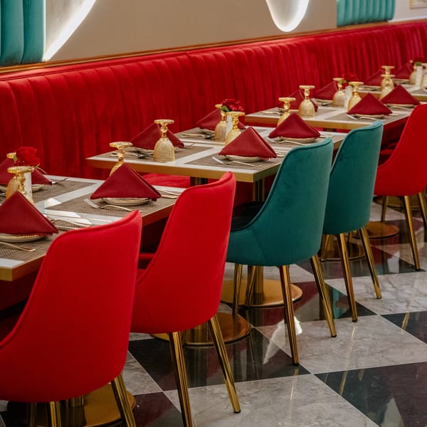 Restaurant interior with red booth seating, teal and red chairs, and set tables with gold accents.