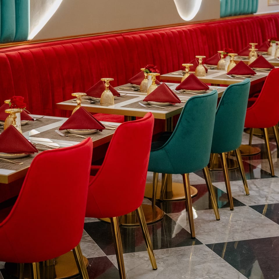 Restaurant interior with red booth seating, teal and red chairs, and set tables with gold accents.