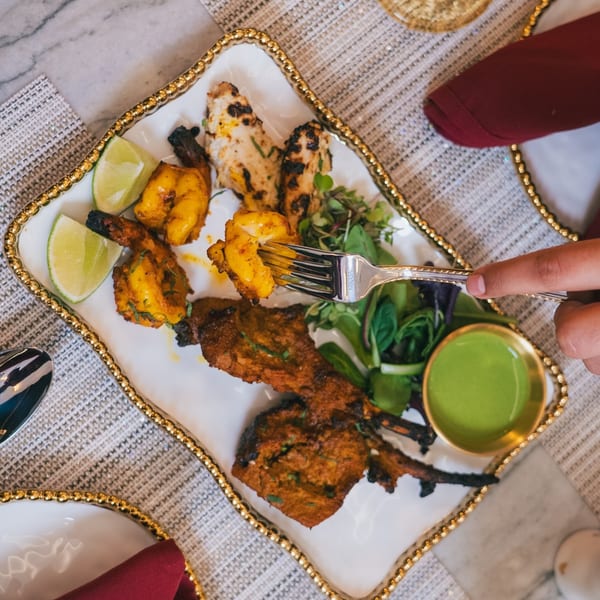 A person's hand uses a fork to pick up a piece of grilled shrimp from a platter of Indian food.