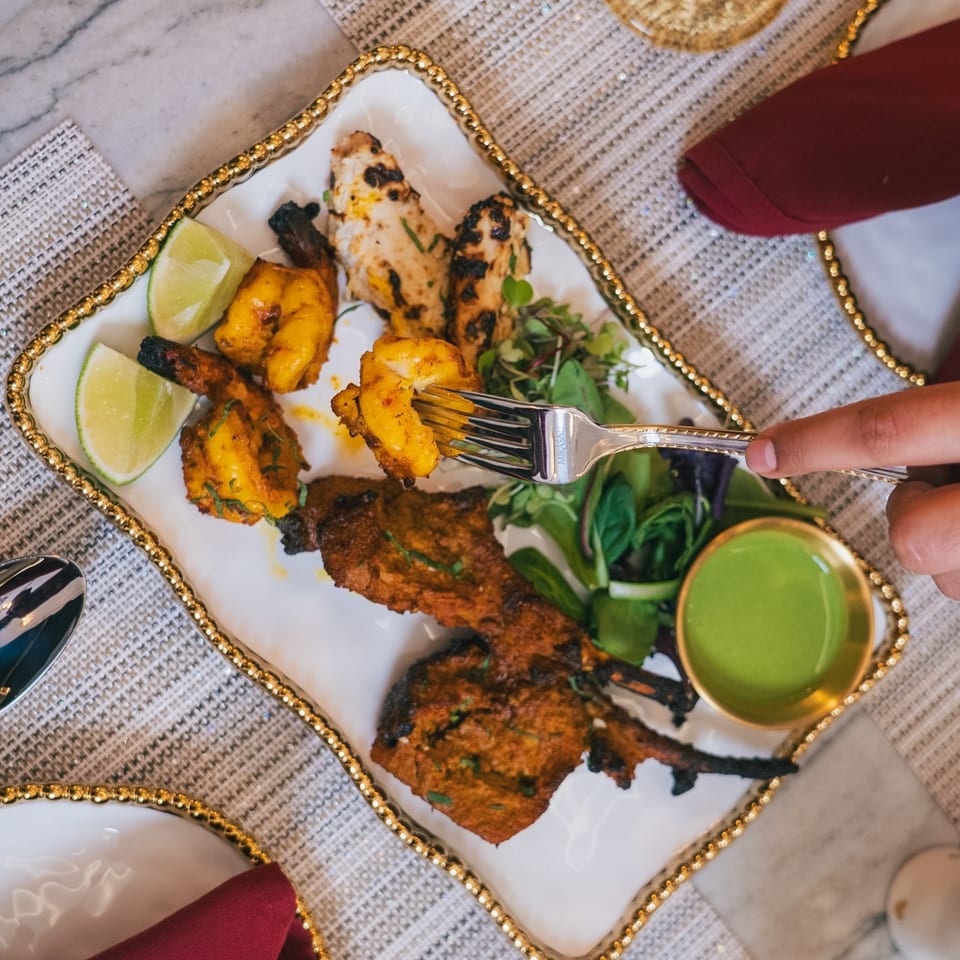 A person's hand uses a fork to pick up a piece of grilled shrimp from a platter of Indian food.